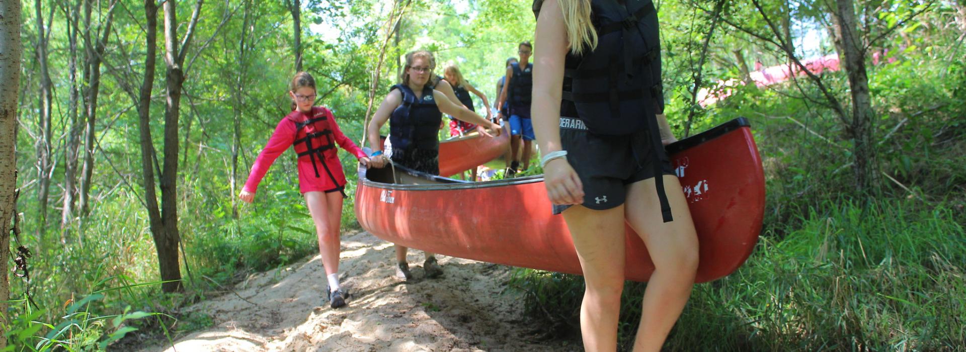 A staff member and two campers carry a canoe down a path