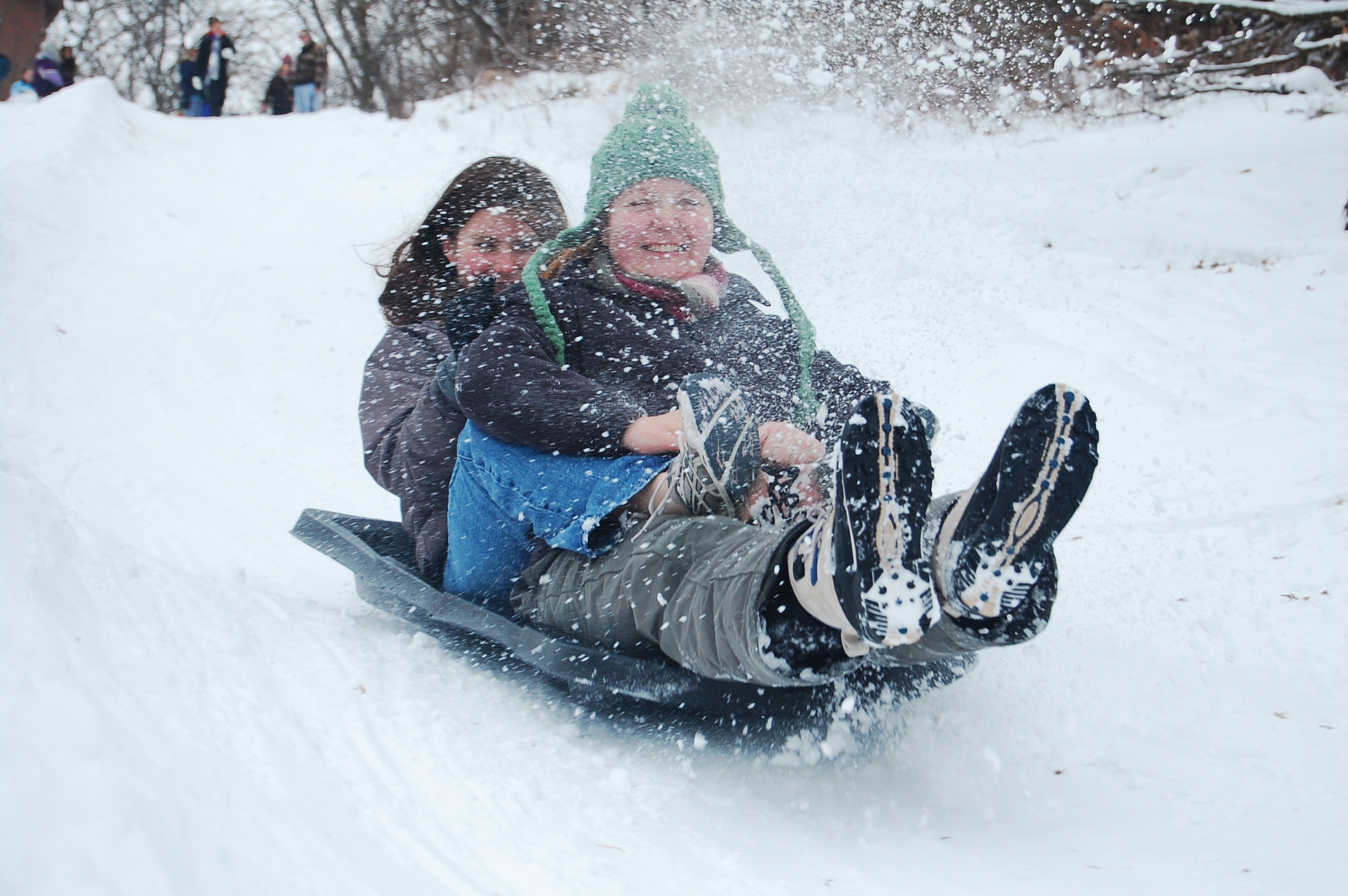 Two people ride a sled down hill as snow is kicked up.