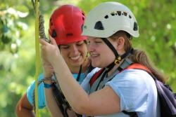 A camper smiles as she is set up to go off the zipline by a staff member