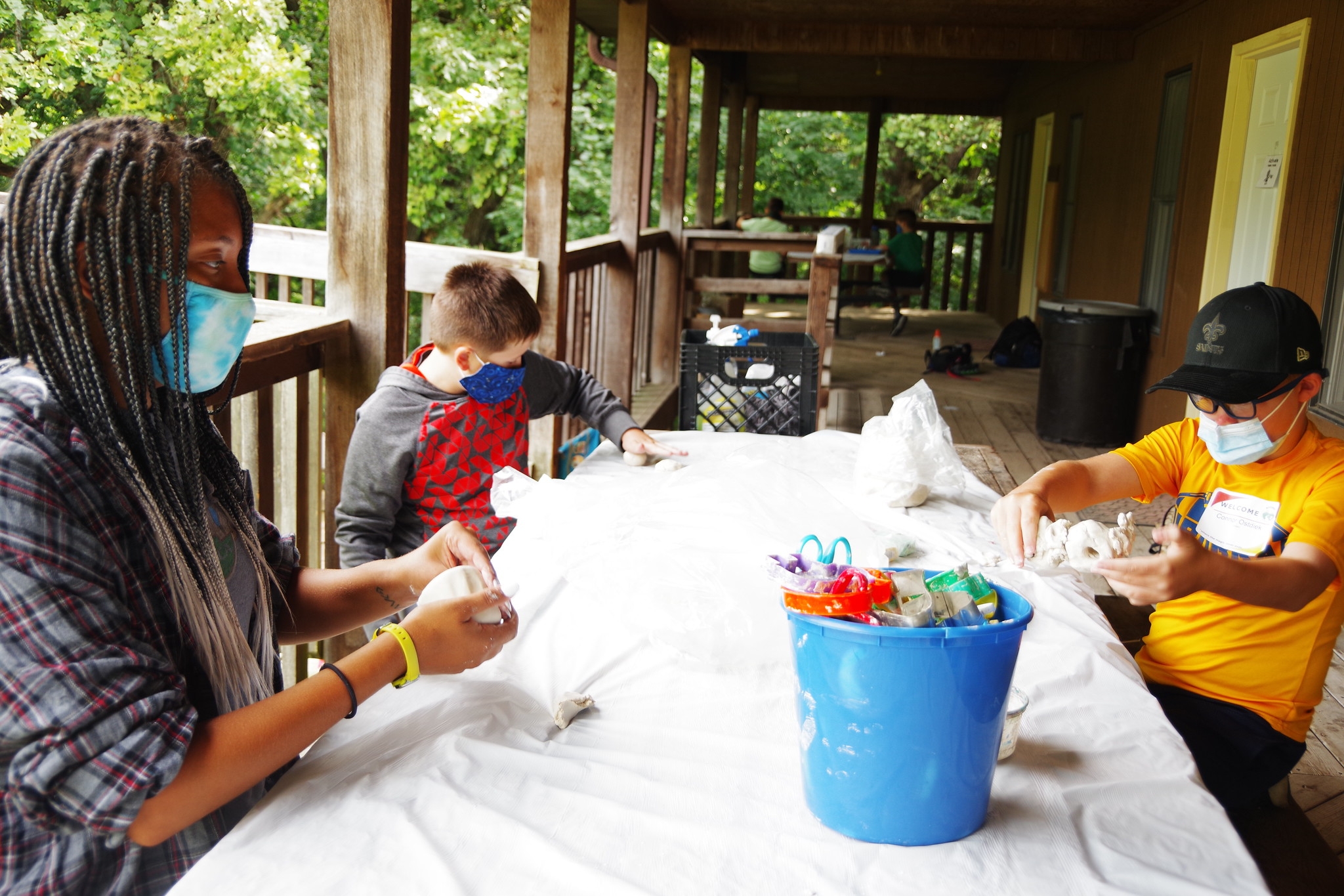 A counselor and two campers play with clay outside while wearing masks