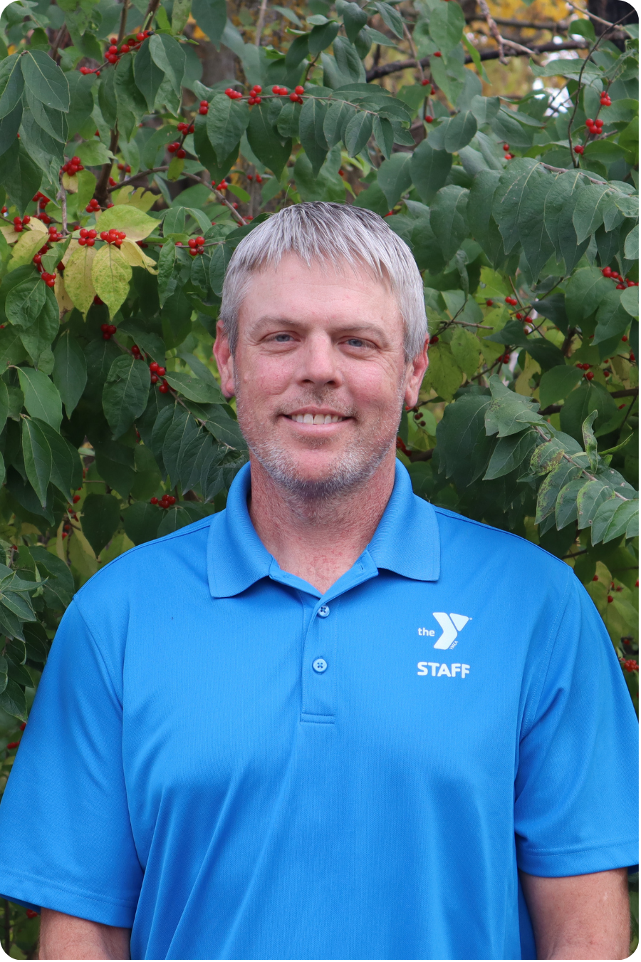 Andrew stands smiling in front of some greenery wearing a blue polo