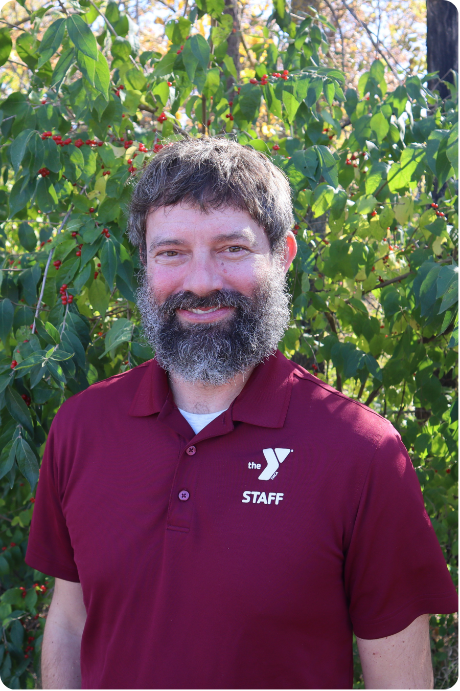 Jason, the director, stands smiling in front of some greenery wearing a maroon polo