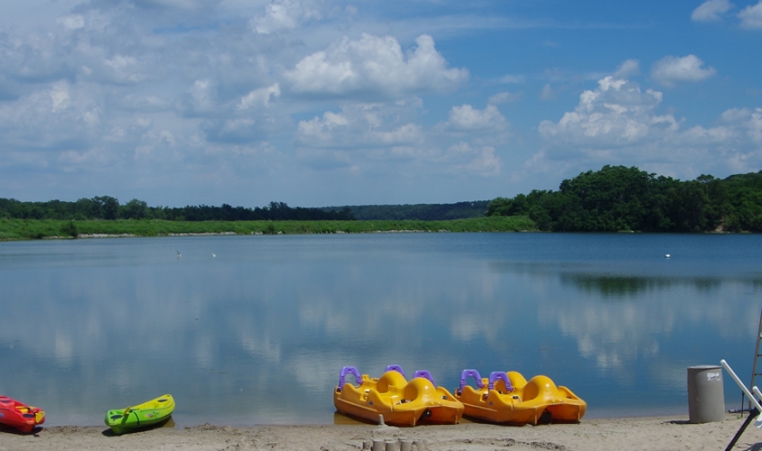 A landscape scene of a lake with canoes, kayaks and paddle boats on the beach
