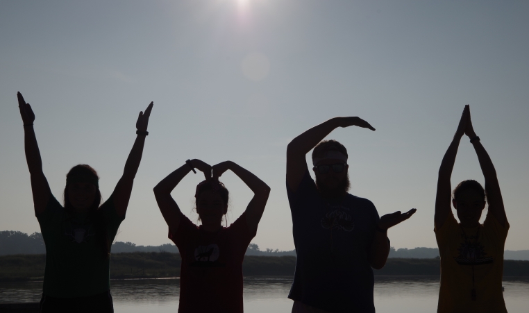 Four people make the letters YMCA with their arms as silhouettes in front of the camp lake
