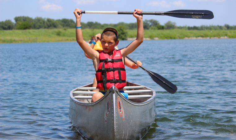 A camper sits at the front of a canoe and holds his paddle over his head in triumph