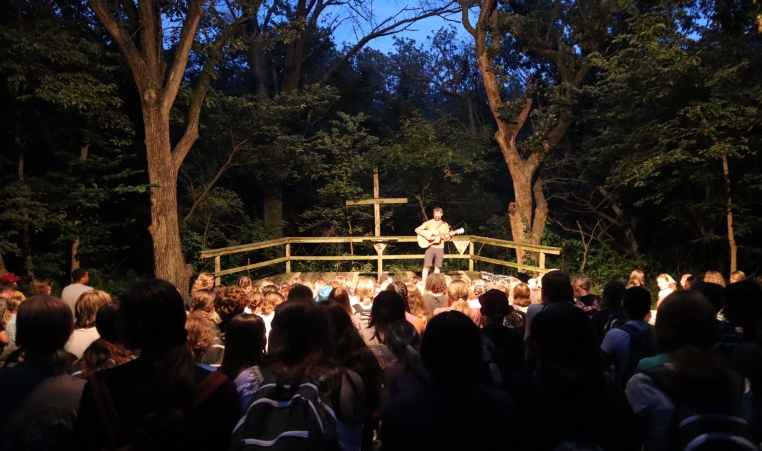 A person stands on stage with a guitar in a wooded amphitheater
