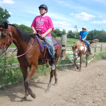 Three campers ride horses along the arena fence line under a blue sky