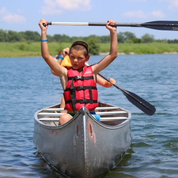 A camper sits at the front of a canoe and holds his paddle over his head in triumph