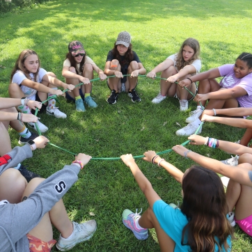 A group of kids sit in a circle leaning back against a loop of rope using it to stand as a team at camp.  