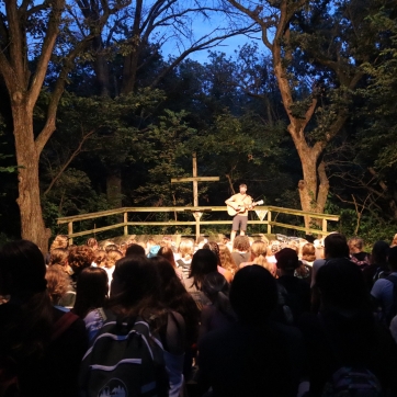 A person stands on stage with a guitar in a wooded amphitheater