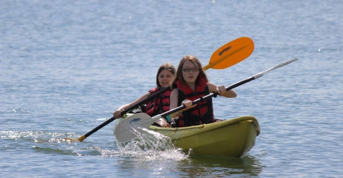 Two girls paddle a sit on top kayak on a lake.
