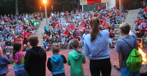 a line of campers with their backs to the camera sing a song in front of the camp audience which is visible in the background.