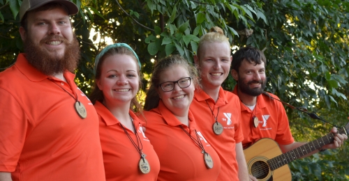 A line of smiling staff in orange polos are lined up, arm in arm. One is holding a guitar. 