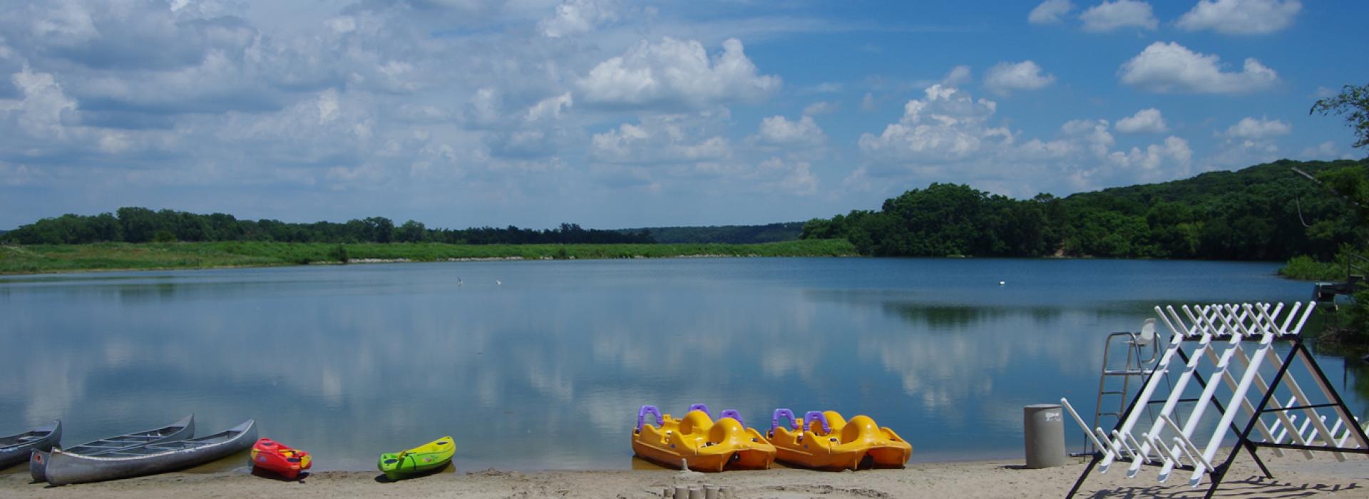 A landscape scene of a lake with canoes, kayaks and paddle boats on the beach
