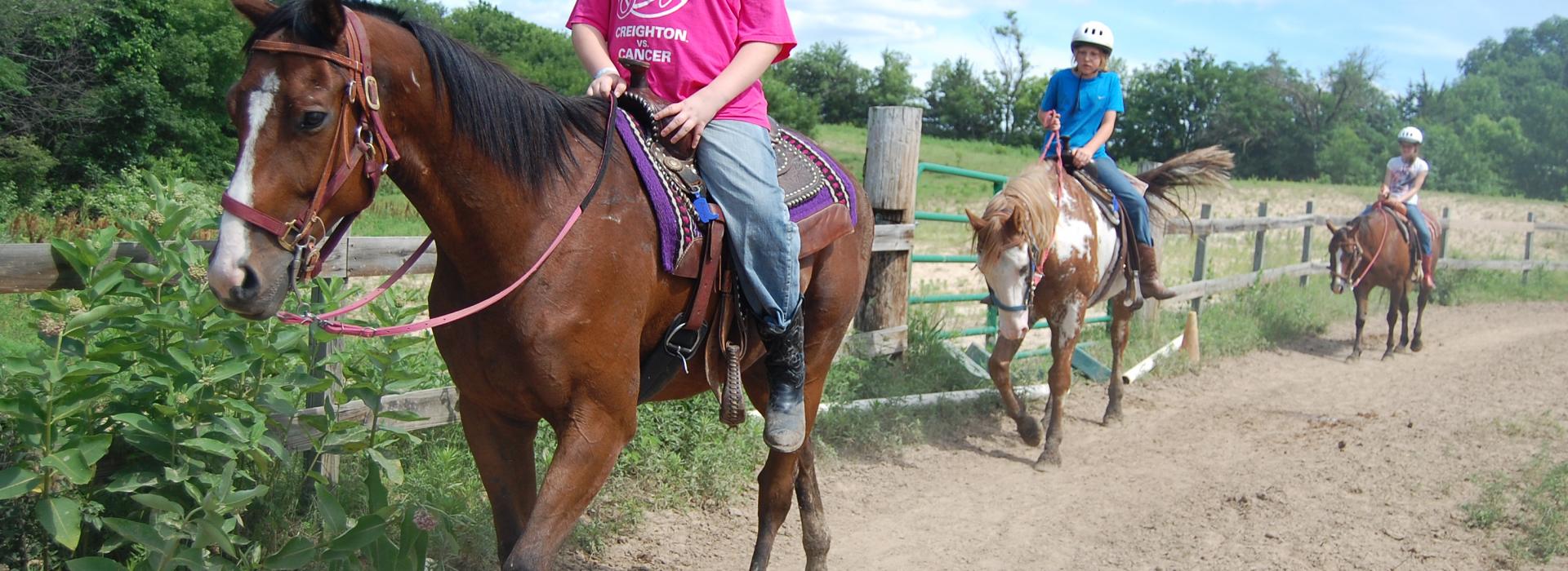 Three kids ride horses along an arena fence line under a blue sky with clouds.