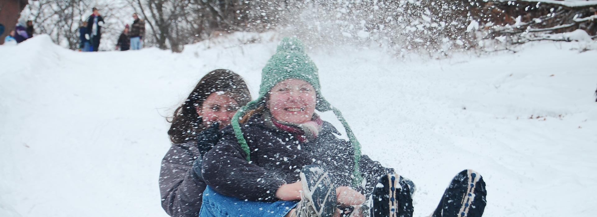 Two people ride a sled down hill as snow is kicked up.