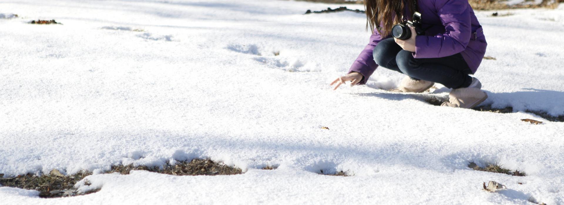A camper kneels in the snow to draw a picture while holding a camera