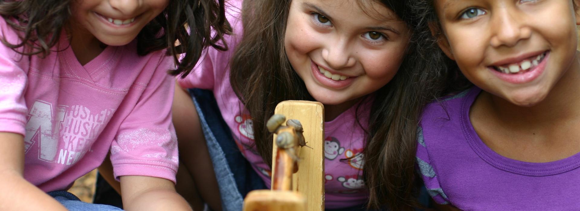 Three girls smile as they look at the handle of a bucket that has three snails they collected on it.