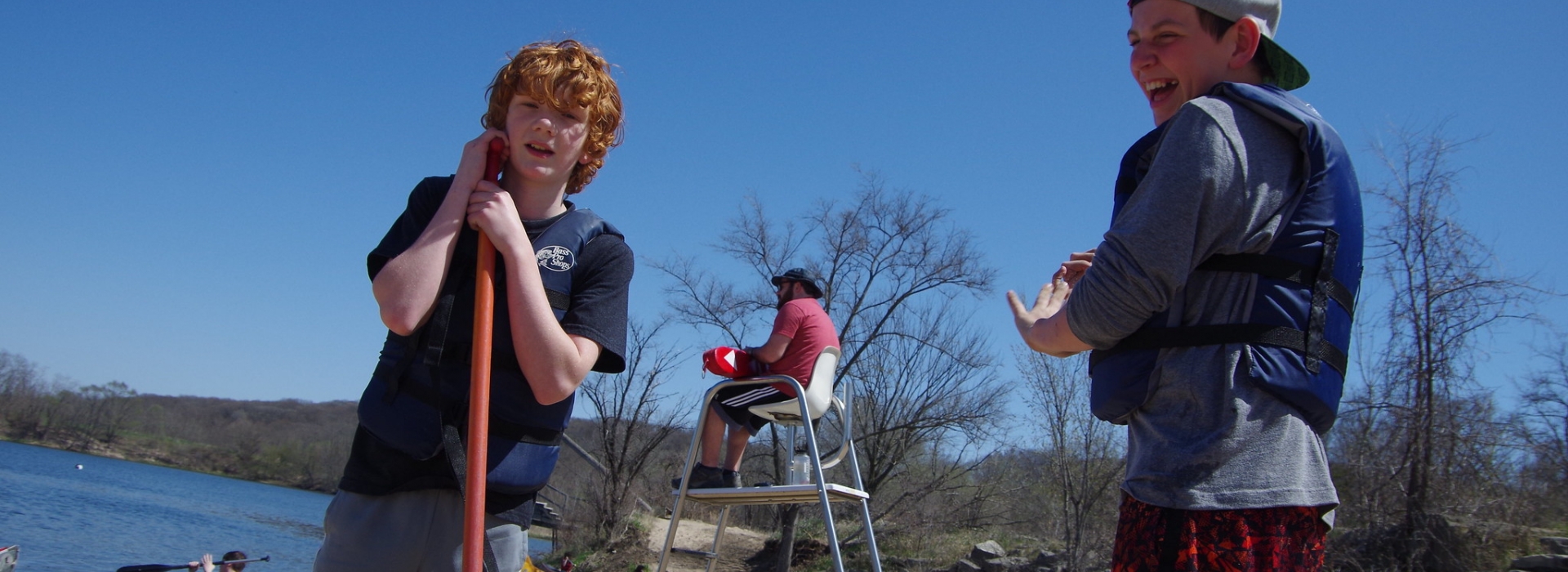 Two campers in life jackets get ready to canoe with the lifeguard in the background