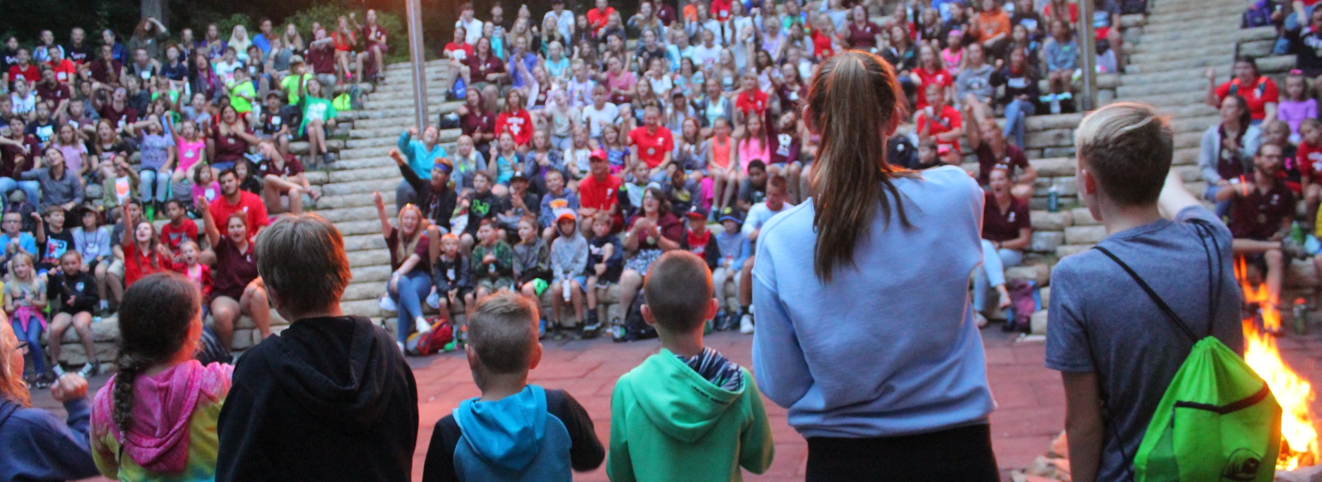 a line of campers with their backs to the camera sing a song in front of the camp audience which is visible in the background.