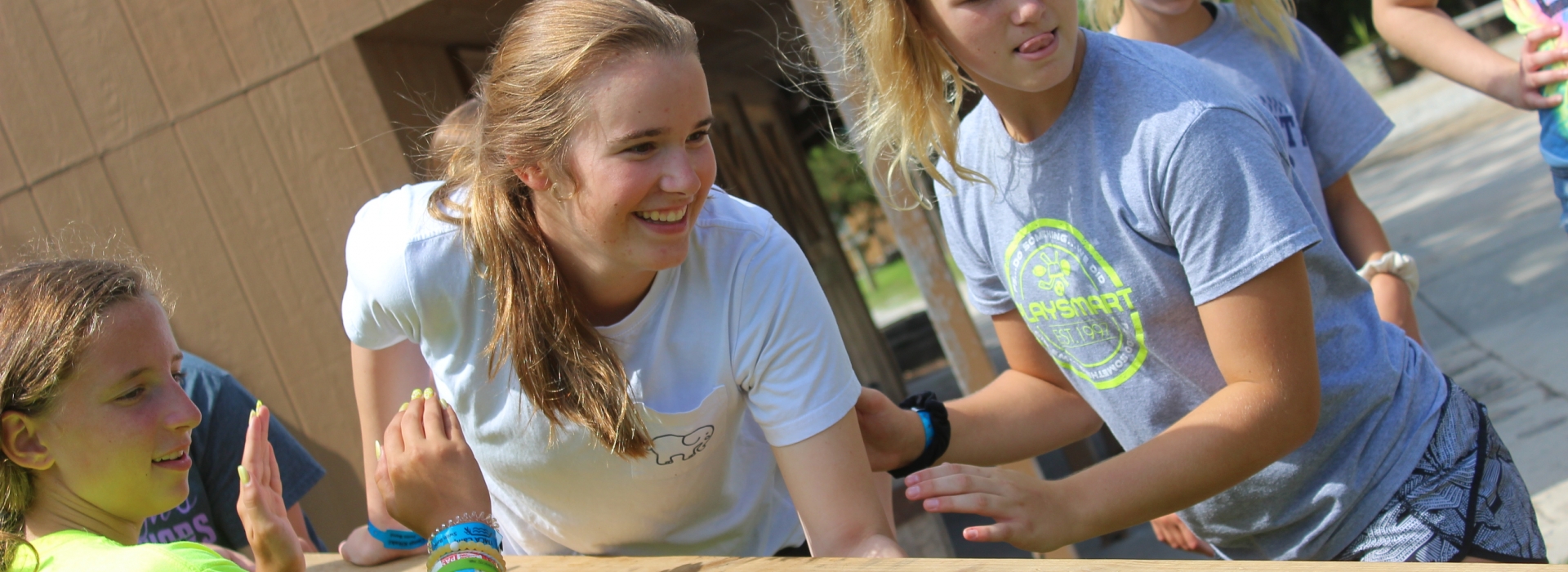 A camper holds a board and leans forward to put it in place while two cabin mates spot her to keep her safe