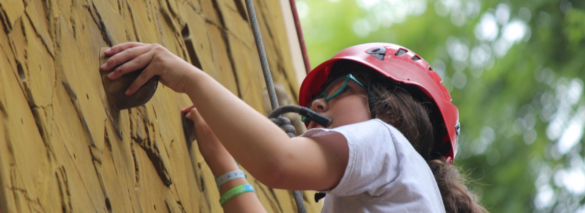 A camper with helmet and harness holds a climbing rock as she scales the Tango Tower