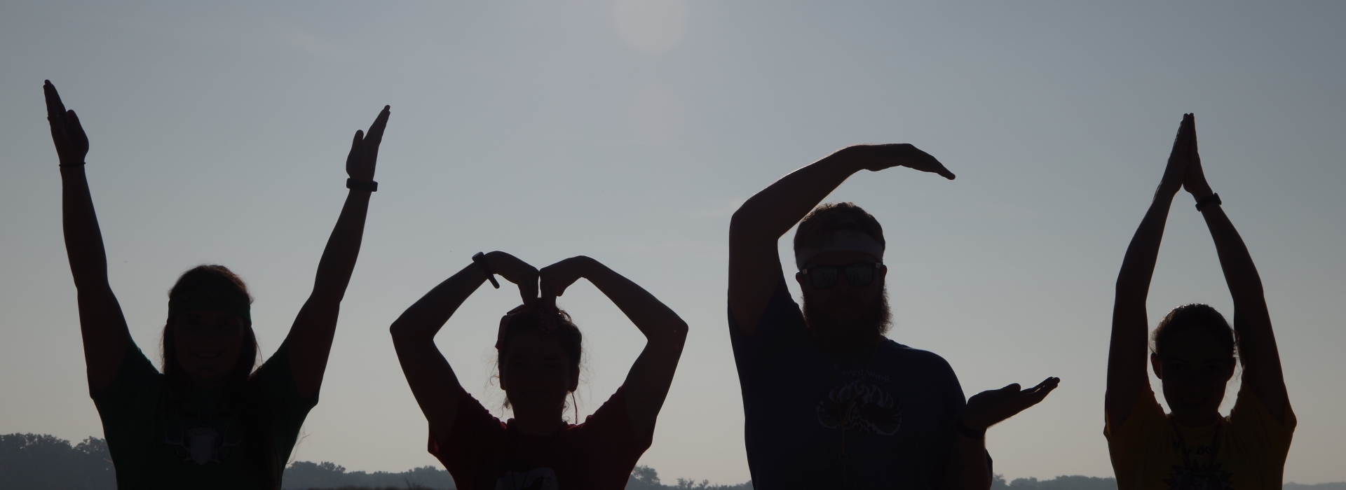 Four people make the letters YMCA with their arms as silhouettes in front of the camp lake