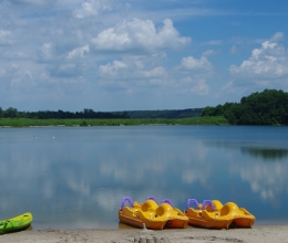 A landscape scene of a lake with canoes, kayaks and paddle boats on the beach