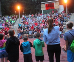 a line of campers with their backs to the camera sing a song in front of the camp audience which is visible in the background.